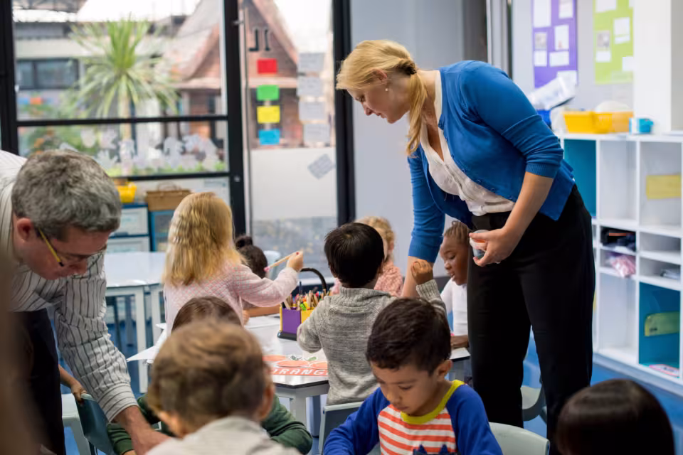 Eine Klassenassistenz geht von Tisch zu Tisch und unterstützt die Kinder bei einer Aufgabe.