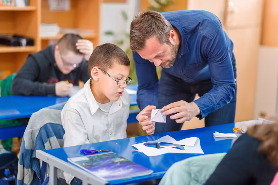 Ein Schulbegleiter hilft einem Schüler beim Falten einer geometrischen Form.